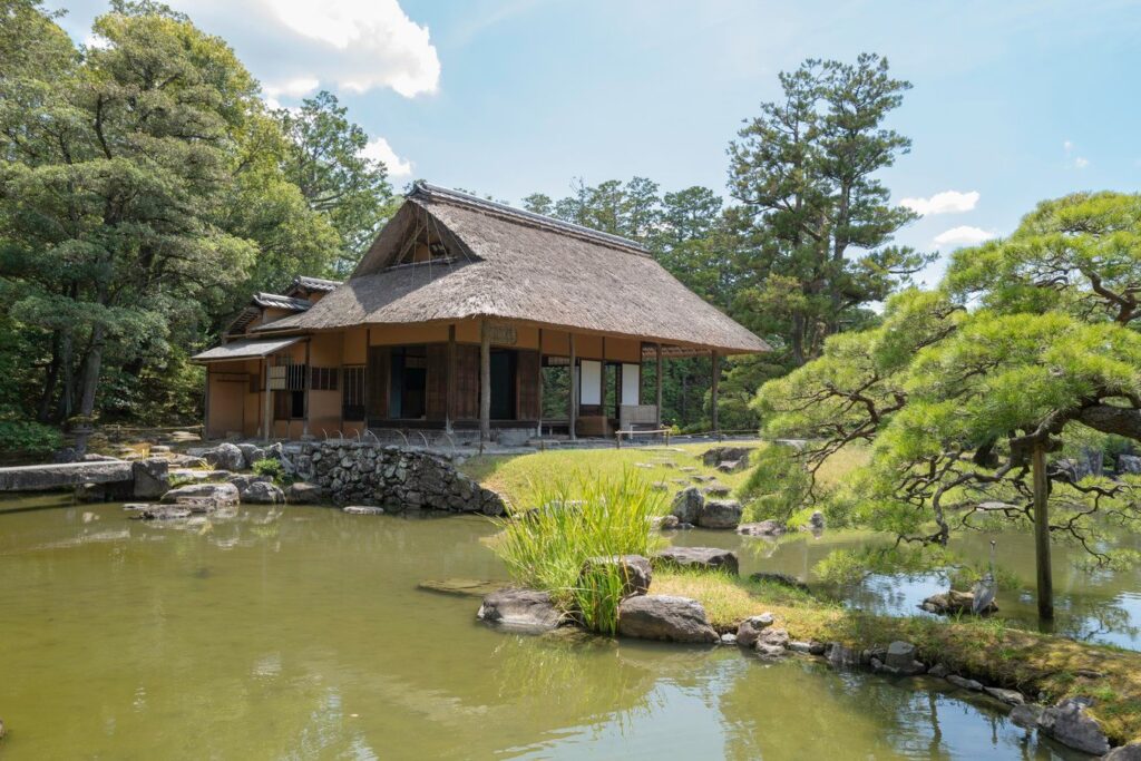 Traditional Japanese thatched-roof house by a serene pond, surrounded by lush greenery. Perfect for Japan tour explorations.