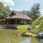 Traditional Japanese thatched-roof house by a serene pond, surrounded by lush greenery. Perfect for Japan tour explorations.